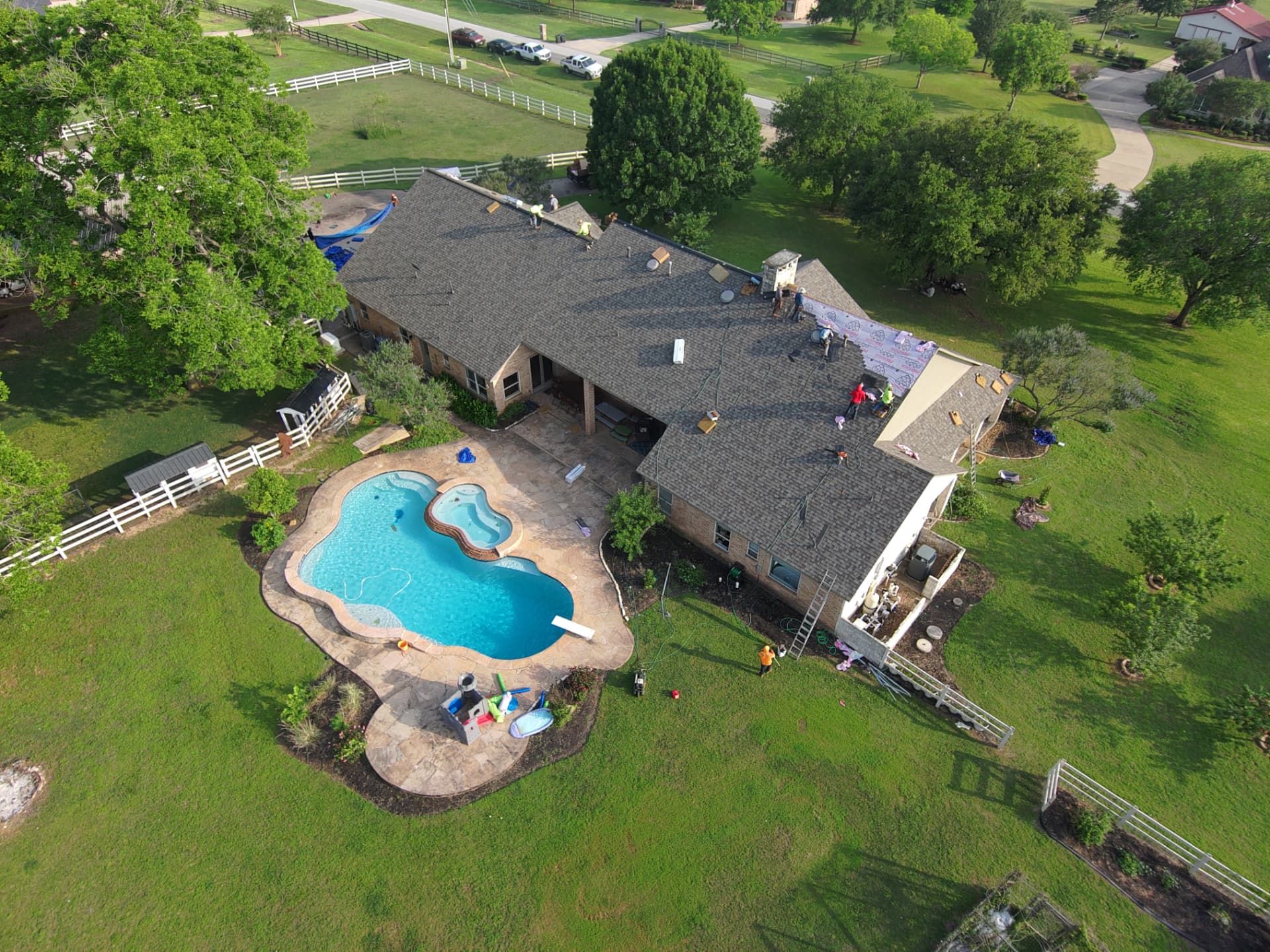 Aerial drone photo of crews installing new shingles on a ranch-style home with pool in Fort Bend County